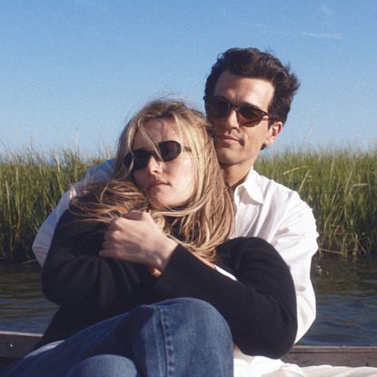 A blonde woman (Sarah Pidgeon as Carolyn Bassette Kennedy) and a brunette man (Paul Anthony Kelly as JFK Jr.), both wearing sunglasses, cuddle while sitting in a small boat on the water. A shoreline covered in reeds is seen in the background, in a still from FX's 'Love Story.'